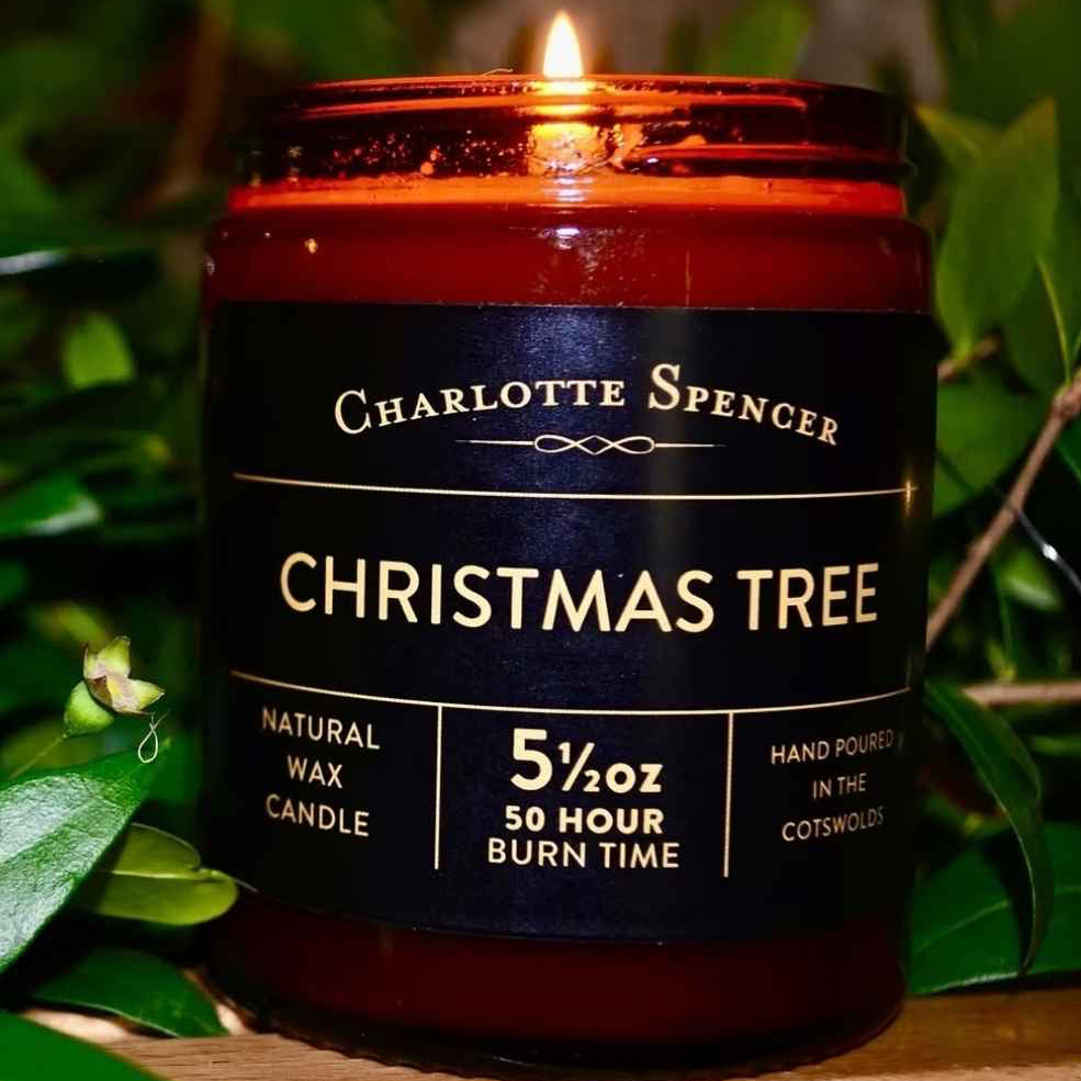 A Charlotte Spencer Christmas Tree fragranced candle in an amber glass jar with black label and green foliage in the background.